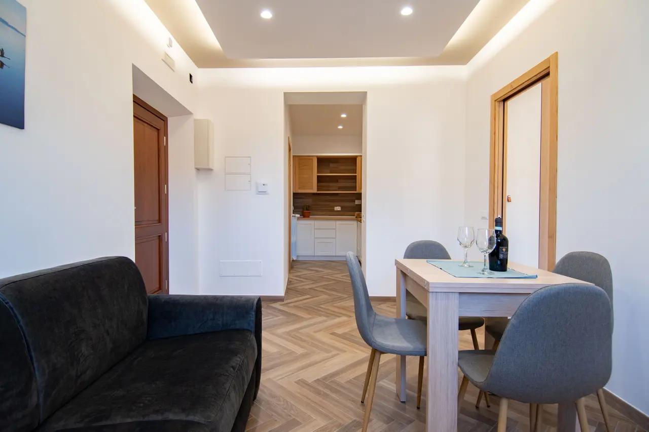 Zona living luminosa con divano e tavolo da pranzo, collegata alla cucina in un ambiente armonioso e ben organizzato. Il divano, all’occorrenza, si trasforma in